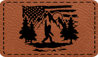 Close-up of the 'Bigfoot Flag Outdoors' leatherette patch featuring Bigfoot throwing rock horns in front of a mountain range with an American flag backdrop.